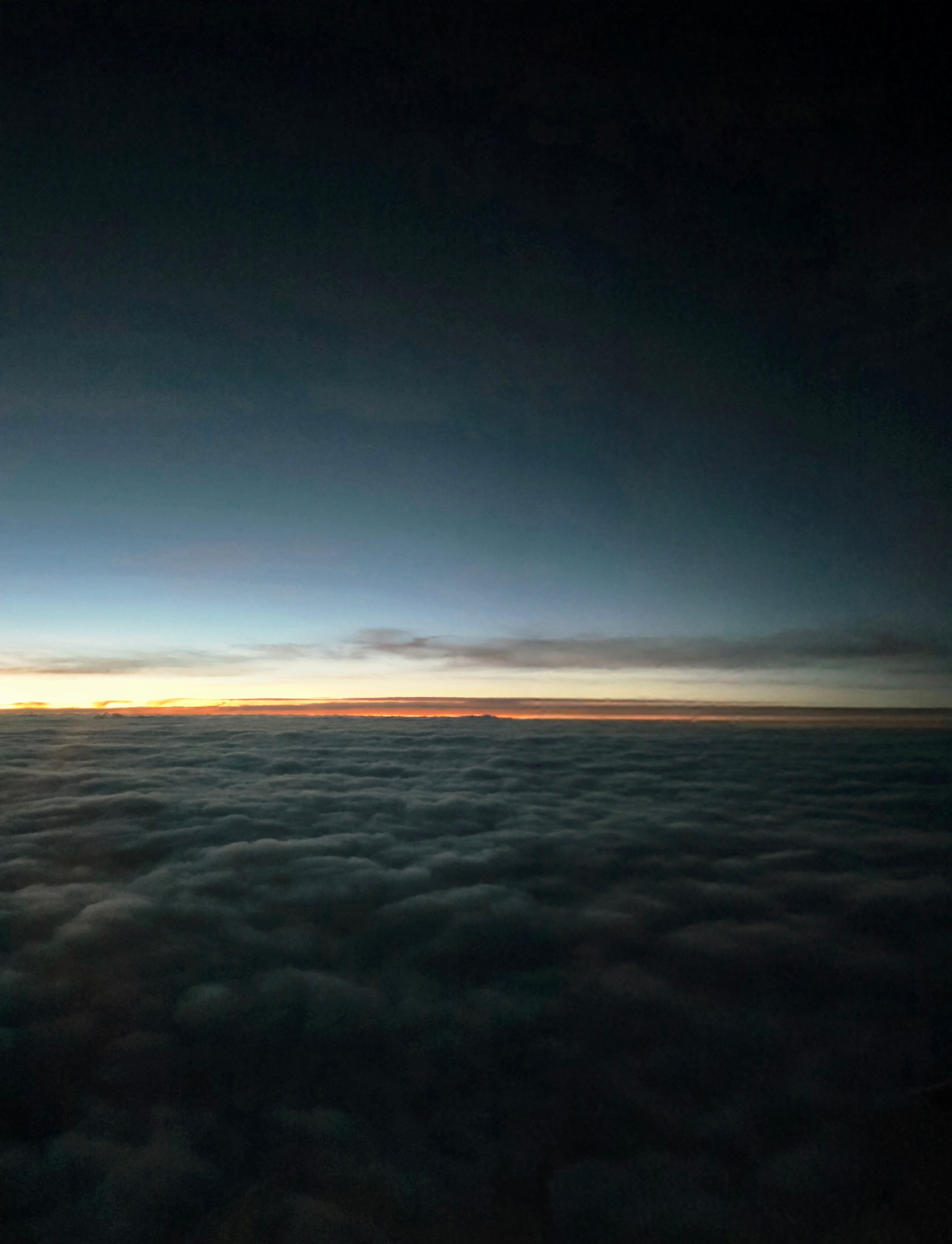 View from an airplane window above a thick layer of clouds at dusk. A thin orange line of sunset glows along the horizon under a dark blue sky.