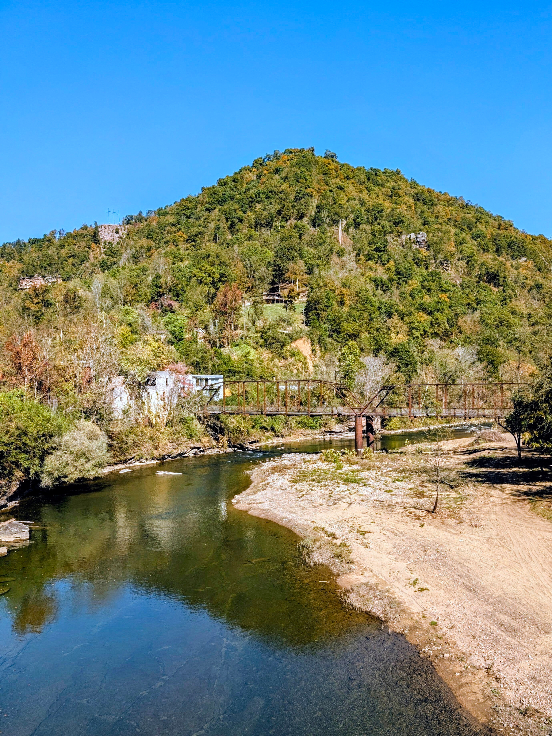 Auto-generated description: A lush, green hill rises behind a narrow river with a rusty bridge crossing it under a clear blue sky.
